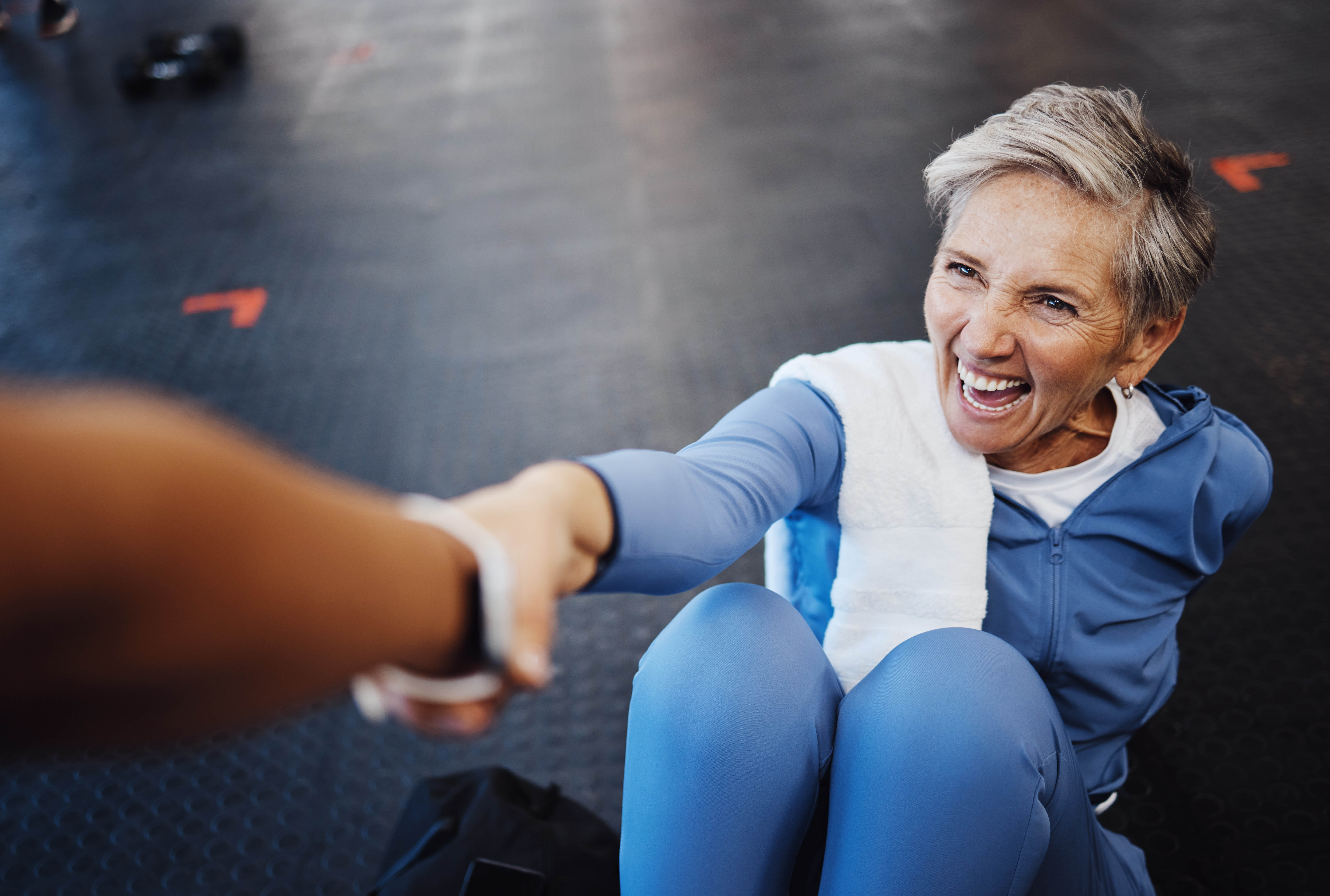 Older woman laying on the floor in the gym reaching up towards a hand that is reaching to pull her up